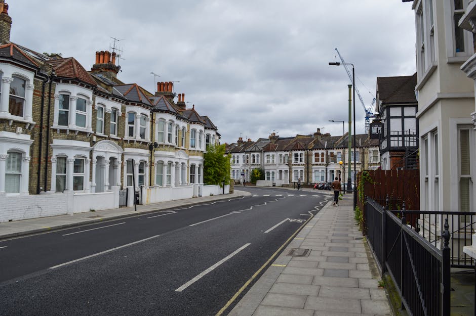 A quiet residential street in Hendon showing a row of Victorian-style terraced houses with bay windows, brick facades, and tiled roofs. The pavement runs parallel to the road, which has parked cars along the curb. Overcast skies cast diffuse light over the scene, with a few pedestrians walking along the sidewalk. In the foreground, there is a black metal fence bordering one property, and a small green shrub is visible next to it. The street is lined with traditional street lamps and a crane can be seen in the background, indicating ongoing construction or maintenance work. This setting reflects typical London residential architecture and environment, suitable for a home relocation or furniture transport process, as handled by [COMPANY_NAME], providing professional removals and moving services in Hendon. The scene emphasizes the urban infrastructure and logistical considerations involved in local moving and packing operations. 