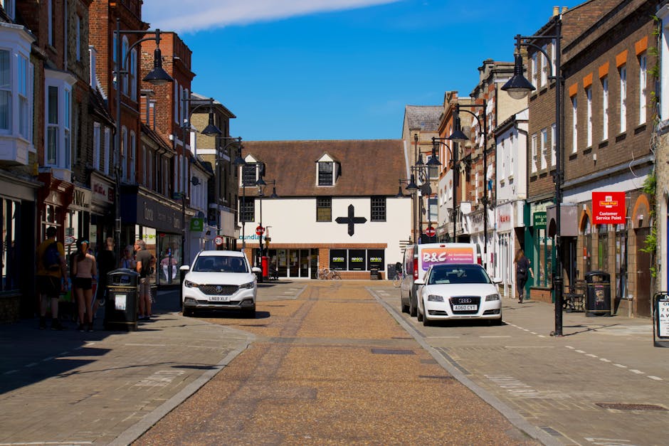 A street scene in Hendon with a clear blue sky, featuring a central pedestrian zone paved with cobblestones and flanked by multi-storey shops and buildings. On the left, a group of people, including some wearing hats and casual clothing, stand near shop entrances, while on the right, a man walks past a supermarket with a red customer service post sign attached to a lamppost. Parked vehicles, including a white van and a white car, are positioned along the pavement, likely part of a home relocation or furniture transport operation managed by Man with Van Hendon. In the background, a white building with a distinctive black cross symbol on its upper façade is visible, indicating a commercial or retail location, with additional street lamps and signage. This scene captures the everyday environment where house removals and packing and moving logistics occur, with vehicles and equipment positioned to facilitate efficient furniture transport and loading processes.