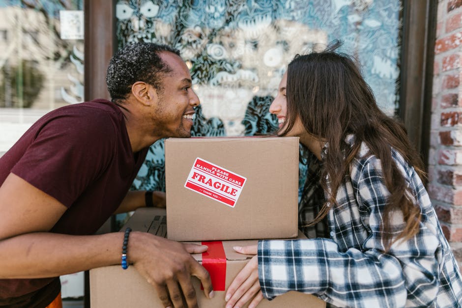 A man and a woman are positioned face-to-face outside a building, smiling at each other while holding and passing stacked cardboard boxes during a home relocation. The man, with short, curly dark hair and a dark red T-shirt, is on the left, and the woman, with long, dark brown hair and a blue, white, and black plaid shirt, is on the right. Between them is a medium-sized cardboard box with a red and white 'Fragile' handling label affixed to it, indicating careful packing and moving processes. Behind them, a large window with patterned curtains is visible, and the scene is set in natural daylight. The image captures a moment of teamwork during furniture transport, with the boxes likely prepared for loading into a van or staged inside the property for packing and moving, highlighting the essentials of professional removals as offered by Man with Van Hendon.