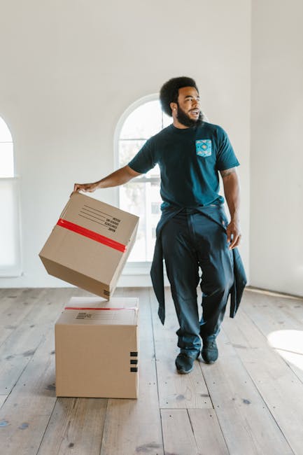 A man with a dark complexion, curly black hair, and a beard is inside an unfurnished room with white walls and wooden floorboards. He is wearing a navy blue t-shirt with a logo on the chest, dark trousers, and has a sweater tied around his waist. He is holding a medium-sized cardboard box with a red stripe and tape sealing it in his right hand, while standing near two other boxes placed on the floor, one of which appears larger and is sealed with black tape. The boxes are ordinary packing cartons likely used for a home relocation process. Behind him, a large arched window with clear glass allows natural light to fill the space, illustrating a typical interior scene during moving and packing activities. The scene supports the context of furniture transport and unpacking as part of house removals, with Man with Van Hendon providing professional moving services. This visual captures the typical steps in packing and loading as part of a professional removals operation in Hendon, London.