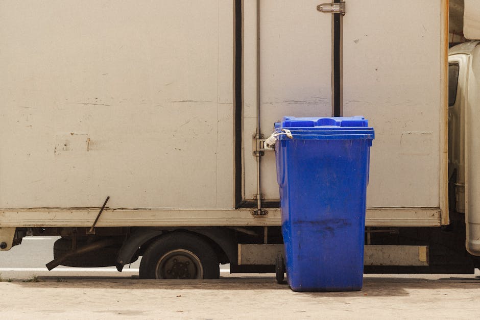A blue recycling bin positioned next to the rear door of a white commercial van during a home relocation or furniture transport process. The van appears to be parked in a driveway or on the street, with the bin placed on the pavement close to the loading area. The van’s side panel shows signs of wear, and part of the vehicle’s wheel and undercarriage are visible beneath the body. The environment is well-lit, likely daylight, and the scene suggests a typical setting for loading or unloading household items or waste during a moving service. Man with Van Hendon utilizes such vehicles and bins in their removals and packing services to assist clients with house moves or disposal of bulky waste, such as furniture or mattresses, in accordance with disposal rules for Hendon. The image underscores the logistics involved in moving or waste collection, with clear focus on the bin and van as part of the relocation process.