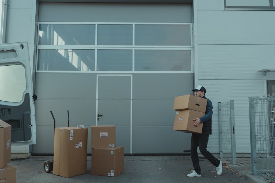 A man wearing a black jacket, dark pants, a black and white cap, and white sneakers is seen carrying two cardboard moving boxes, one stacked on top of the other, during a home relocation process. He i