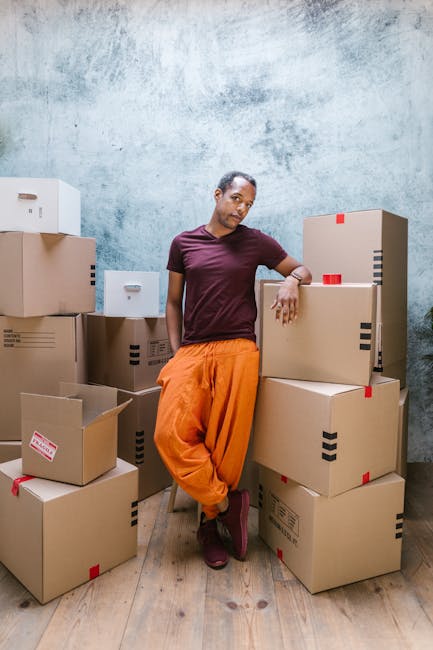 A person standing indoors beside a collection of cardboard boxes of various sizes, some with red tape and labels, stacked on a wooden floor against a textured blue-grey wall. The individual is leaning casually on one of the boxes with their right arm, wearing a maroon T-shirt, orange loose-fitting trousers, and dark shoes. The boxes are arranged around them, suggesting an ongoing home relocation or packing process, typical of furniture transport and moving services. The setting appears to be a designated packing or storage area within a property, with natural or soft lighting illuminating the scene. This image, associated with Man with Van Hendon, visually reflects the logistical aspect of home removals, including packing and loading, in preparation for furniture transport and house moves, aligning with services offered by Hendon removals companies.