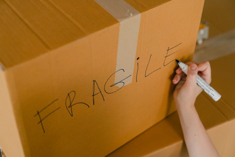 A person’s hand is holding a black marker and writing the word 'FRAGILE' in large, uppercase letters on a cardboard box, which is sealed with clear packing tape. The cardboard box is situated among other similar boxes, indicating a home relocation or packing process. The scene takes place indoors, likely in a room prepared for moving, with wooden flooring visible. The image highlights the labeling process during packing and moving, conducted by Man with Van Hendon, a company specializing in removals and furniture transport services. The careful labeling of fragile items ensures cautious handling during the loading process into a removal van, exemplifying professional packing practices for house removals and minimizing the risk of damage during transport.