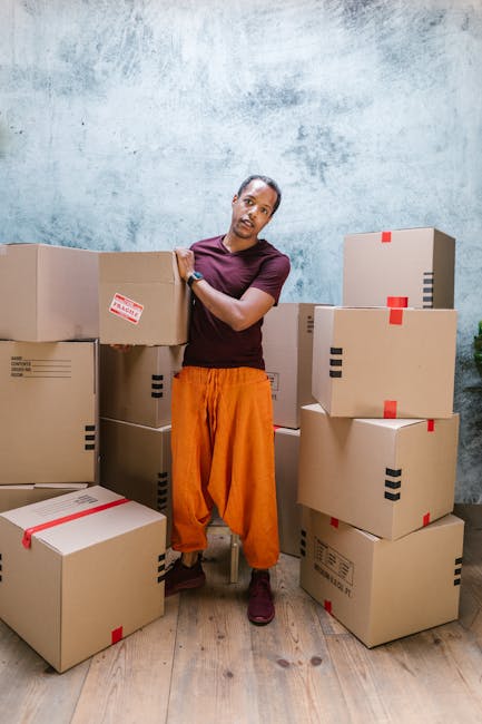 A person standing indoors beside a collection of cardboard boxes of various sizes, some with red tape and labels, stacked on a wooden floor against a textured blue-grey wall. The individual is leaning casually on one of the boxes with their right arm, wearing a maroon T-shirt, orange loose-fitting trousers, and dark shoes. The boxes are arranged around them, suggesting an ongoing home relocation or packing process, typical of furniture transport and moving services. The setting appears to be a designated packing or storage area within a property, with natural or soft lighting illuminating the scene. This image, associated with Man with Van Hendon, visually reflects the logistical aspect of home removals, including packing and loading, in preparation for furniture transport and house moves, aligning with services offered by Hendon removals companies.