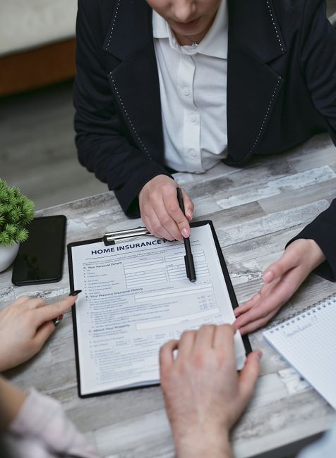 A person wearing glasses and a checked blazer is seated at a wooden table, holding a clipboard with a printed insurance and liability form related to house removals in Hendon, as part of a home reloca