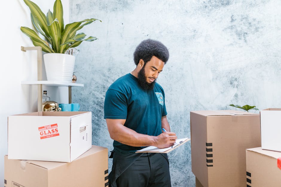 A man with a curly afro hairstyle and a full beard wearing a blue T-shirt is inside a room, standing among stacked cardboard boxes used for packing during a home relocation. The boxes are of various s