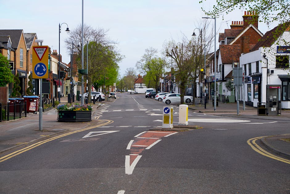 A wide view of a narrow street on The Burroughs in Hendon, showing typical residential and commercial buildings with brick facades, pitched roofs, and shopfronts. The street features parked cars along both sides, with a central traffic island containing a roundabout sign. Street signs and lampposts line the pavement, and small trees with budding green leaves are visible, suggesting early spring. The road markings include double yellow lines on the sides, directional arrows, and a red-bricked pedestrian crossing section, indicating a restricted-access area. This setting illustrates the challenges of home relocation or furniture transport in tight urban environments, with the scene captured during daylight hours under overcast skies. Man with Van Hendon occasionally supports such relocations, focusing on navigating narrow street access and careful loading processes for effective removals.