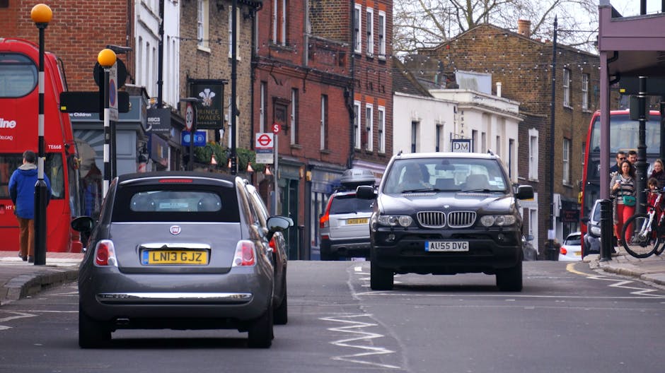 A view of a narrow city street with parked cars on either side, including a grey Fiat 500 and a black BMW SUV, both situated within designated parking bays. The street features a mix of brick and stucco buildings, with shopfronts and pedestrians visible on the sidewalks. One sidewalk has a red double-decker bus stop shelter, and there are people waiting and walking nearby, some with bicycles. The scene is set during daytime with overcast weather, and the street appears busy yet orderly. This environment illustrates typical urban conditions encountered during home relocation or furniture transport in tight-access areas, often requiring careful moving logistics by companies like Man with Van Hendon, especially on streets like The Burroughs where space is limited for larger vehicles and efficient packing and loading are essential.