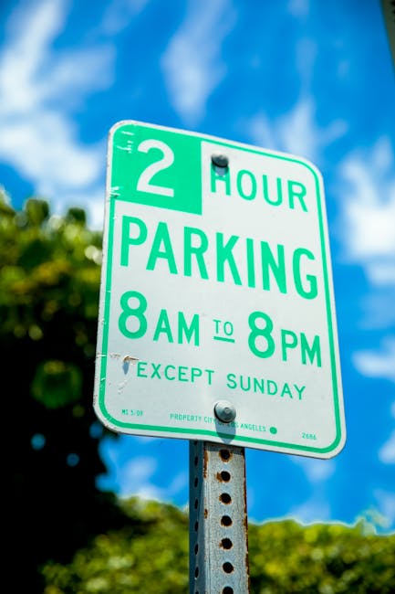 A green and white parking restriction sign mounted on a metal post displays a 2-hour parking limit from 8 am to 8 pm, excluding Sundays, with the notice issued by Los Angeles city authorities. The sign is positioned outdoors against a backdrop of partly cloudy blue sky and blurred green foliage, indicating an urban street environment. This type of parking regulation is relevant to planning home relocations or removals in Hendon, as it informs movers of parking restrictions that may affect loading and unloading at the property. Man with Van Hendon, a local removals company, considers such parking signs when organising furniture transport and moving logistics to ensure compliance with local rules and efficient loading processes during house moves, especially in areas with regulated parking hours.
