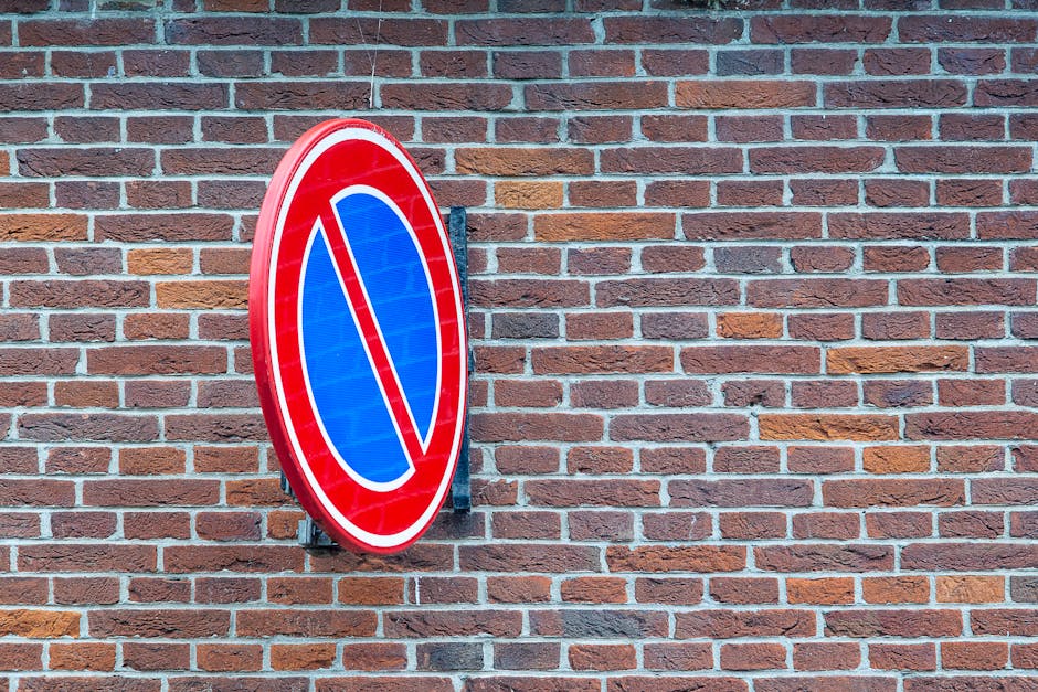 A circular no parking traffic sign is mounted on a black metal bracket affixed to a red brick wall, with the sign featuring a red border, a blue background, and a red diagonal line across it, indicating no parking zone. The brick wall consists of rectangular bricks in shades of red, brown, and tan, arranged in a running bond pattern, with light gray mortar joints. The sign is positioned to the left side of the image, and the wall extends to the right, showing a consistent brick pattern. The lighting appears natural, illuminating both the sign and the textured surface of the brick wall. This setting might be in an urban or residential area, relevant to house removals or moving services, where parking restrictions are often a consideration during home relocation or furniture transport. Man with Van Hendon occasionally operates in such environments, assisting with logistics for house moves and ensuring compliance with parking regulations, as indicated by the proximity of the sign to the building entrance, which is common in property access management during removals.