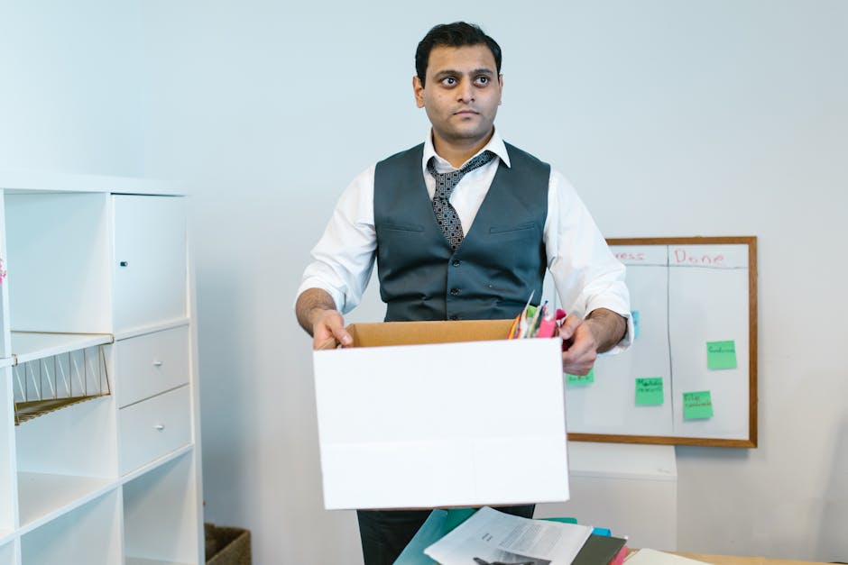 An office worker, dressed in a white shirt with rolled-up sleeves, a dark vest, and a patterned tie, stands inside an office space holding a cardboard box filled with office supplies such as pens, fol