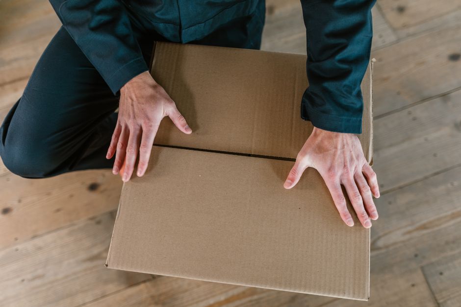 An office worker, dressed in a white shirt with rolled-up sleeves, a dark vest, and a patterned tie, stands inside an office space holding a cardboard box filled with office supplies such as pens, fol