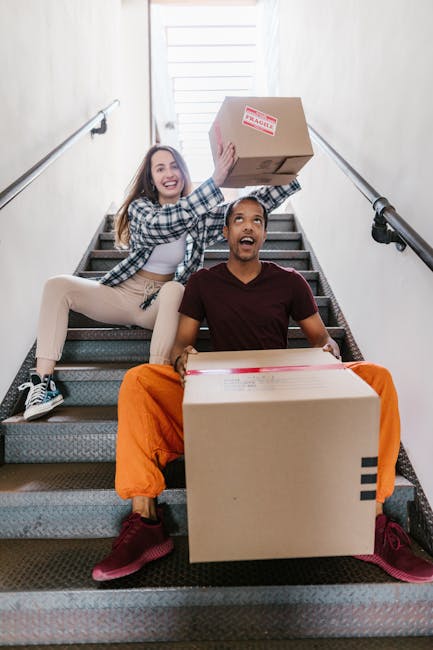 Two young adults, a woman and a man, are sitting on a staircase inside a building, each holding cardboard boxes filled with household items. The woman, positioned on the upper step, is smiling broadly and wearing a checkered shirt, beige pants, and sneakers, while the man, seated below her, is also smiling and dressed in a maroon t-shirt, bright orange pants, and sneakers. They appear to be in the process of packing or unpacking during a home relocation or moving process. The staircase has dark, textured steps with a silver handrail on each side, and natural light illuminates the scene from a doorway or window at the top of the stairs, adding a bright, neutral environment. This setting illustrates the loading or unpacking stage typical of a professional house removals service such as Man with Van Hendon, specializing in timely and efficient moves, including packing and furniture transport.