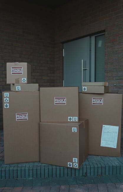 A young woman with curly black hair, dressed in casual clothing and white sneakers, is seated on a wooden floor inside a room surrounded by stacked cardboard boxes of various sizes. Some boxes are sealed with red tape, while others are partially open or labeled with black text and barcodes. She is smiling and resting her head and arms on a large box in front of her, conveying a sense of satisfaction during a home relocation or packing process. The backdrop features a textured, light blue wall, indicating an indoor environment. This scene illustrates the packing and moving activities associated with furniture transport and house removals, which may be handled by services such as Man with Van Hendon, supporting efficient and organized home or office moves.